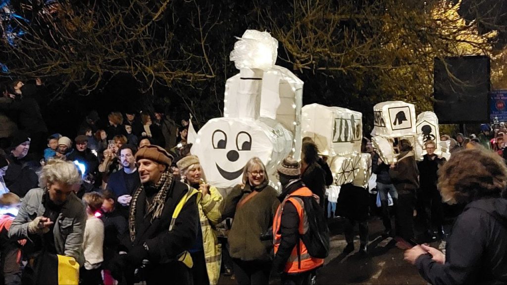 an enormous lantern shaped like a series of train carriages with a smiling engine appears magically to move through a merry, festive crowd